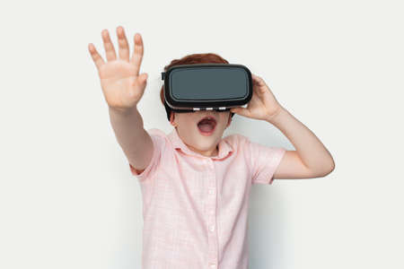 Small caucasian ginger boy wearing a virtual reality headset is touching something on a white studio...