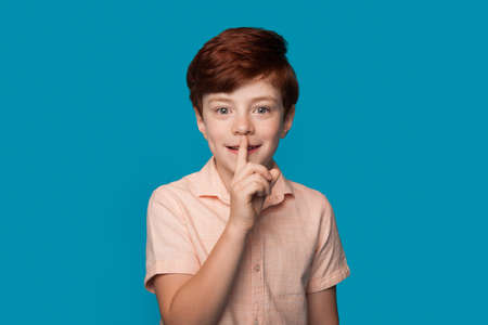 Ginger caucasian boy is gesturing the silence soung smiling on a blue studio wall in casual clothesの写真素材