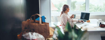 Side view photo of a caucasian senior woman working from home while drinking a coffee and using...