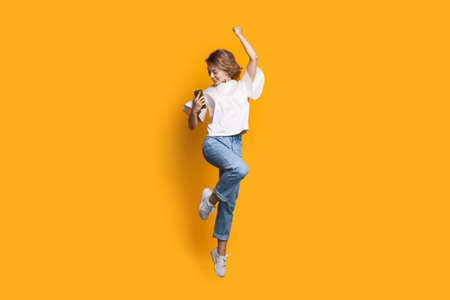 Caucasian woman with blonde hair is gesturing the yes sign on a yellow wall while jumping and...