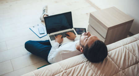 Young caucasian businessman feeling tired after working on the laptop from home sitting on the floor...