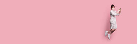 flying caucasian woman in a dress wearing a medical mask is holding a phone and posing on a pink...