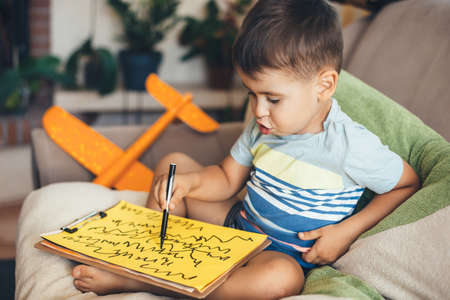 Lovely brunette boy drawing on a yellow paper using a marker while sitting on pillows at home during the international childrens dayの写真素材