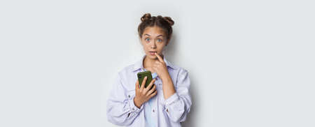 Blonde caucasian lady is gesturing surprisement on a white studio wall by holding a phone and...