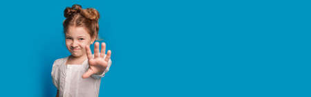 Small caucasian girl is gesturing the stop sign and smile at camera while posing on a blue studio...