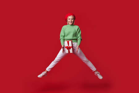 Blonde fashionable woman is jumping on a red wall holding a present box and wear a hat smiling at...