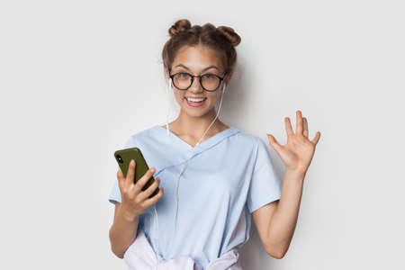 Caucasian woman with blonde hair is smiling while listening to music on a white wall using earphones...