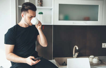 Bearded caucasian man is drinking a coffee while chatting on phone in the kitchen and relaxingの写真素材