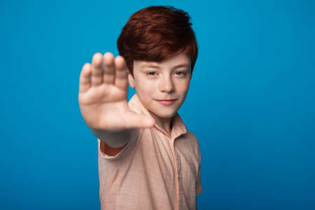 Close up photo of a smiling boy with red hair and freckles gesturing stop sign on a blue wall...