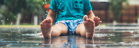 No face photo of a caucasian small boy in blue clothes and barefoot sitting on the ground in water and playingの写真素材