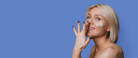 Caucasian woman with blonde hair is applying cream on her face smiling on a blue studio wall with free space having bare shouldersの写真素材