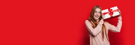 Caucasian woman with red hair and freckles is listening at a present box on a red studio wall with...
