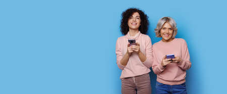 Charming blonde lady with her curly haired friend are chatting on a blue studio wall near free space smiling at cameraの写真素材
