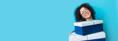 Caucasian curly haired lady is smiling at camera with glasses while holding some present boxes on a...