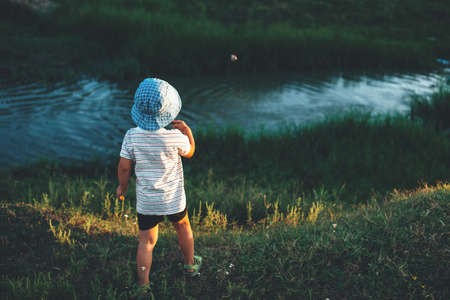 Back view photo of a caucasian boy with blue hat throwing stones in the lake during a summer walk