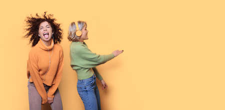 Charming two women dancing on a yellow wall with free space while listening to music