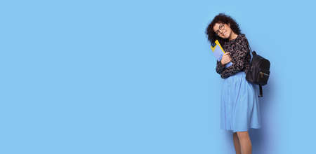 Curly haired student is holding some folders and bag smiling on a blue studio wall with free spaceの写真素材