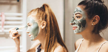 Two caucasian women applying a black mask on their faces looking in the mirror and smileの写真素材