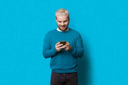 Blonde man with beard is chatting on phone and smile on a blue studio wall