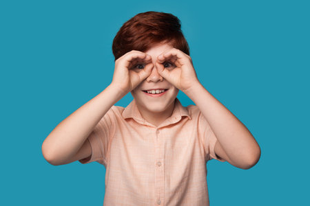 Caucasian ginger boy is gesturing like having binoculars on eyes smiling on a blue background.の写真素材