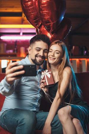 Caucasian couple are making selfie using a phones and smile while holding red balloons on valentines dayの写真素材