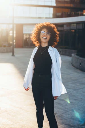 Curly haired business lady with glasses is posing outside and smile at camera during a sunny dayの写真素材