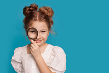 Small girl is covering her eye with a magnifier smiling in a dress at camera on a blue wall with...