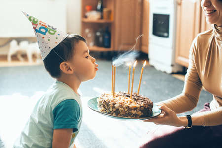 Caucasian mother is holding the cake while son is blowing the candles wearing party hat on his birthdayの写真素材