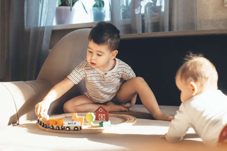 Small caucasian boy is playing on the couch with a toy train railway while newborn sibling is watching himの写真素材
