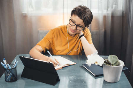 young student with glasses is having online lessons from home using a tablet and writing in a copybookの写真素材