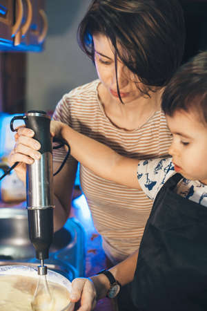 Small caucasian boy is helping his mother preparing food in the kitchen by mixing somethingの写真素材