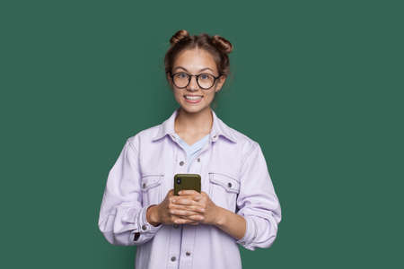 Blonde caucasian girl is smiling toothily at camera on a green wall holding a mobile and wearing glassesの写真素材
