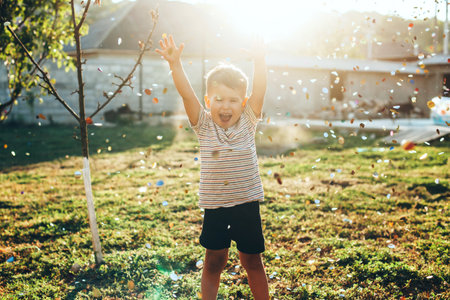 Caucasian boy is playing between a lot of confetti flying near him in the backyard at home on a green fieldの写真素材