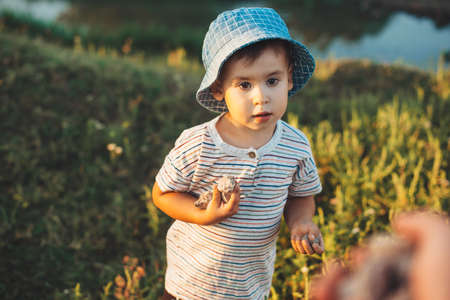 Small caucasian boy with a blue hat is playing near a lake collecting stones while somebody is giving some moreの写真素材