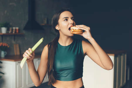 Brunette sporty woman is eating a burger while holding a leek in her handsの写真素材