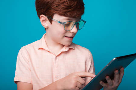 Close up photo of a caucasian ginger boy wearing glasses who is using a tablet on a blue studio wallの写真素材
