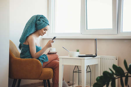 Caucasian lady after taking a bath is chatting on a mobile wearing a towel on head and drinking a teaの写真素材
