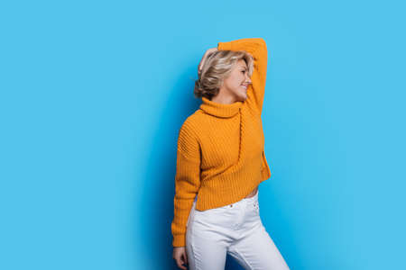 Blonde fashionable woman in a warm sweater is smiling while posing on a blue wall with free spaceの写真素材
