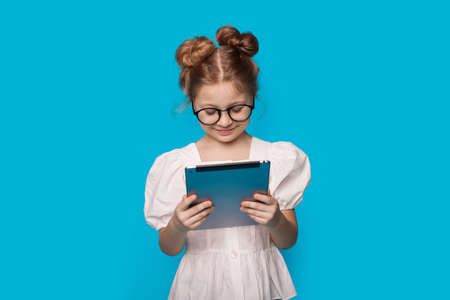 Caucasian cute girl is reading from a tablet smiling on a blue studio wall through glasses in a...