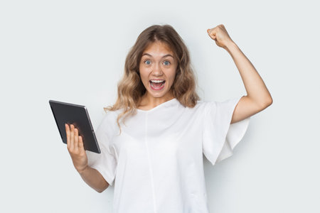Blonde woman is gesturing the yes sign on a white studio wall holding a tablet and smile at cameraの写真素材