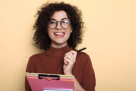 Adorable student with curly hair showing her tongue while holding folder on a yellow studio wall