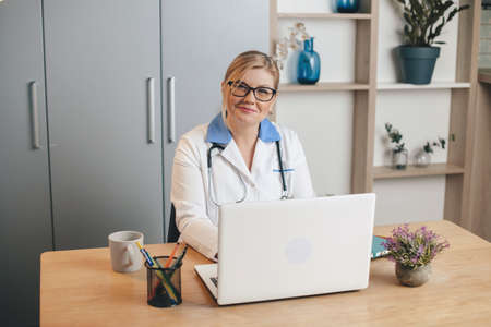 Happy senior blonde doctor smiling at camera while using a laptop at the officeの写真素材