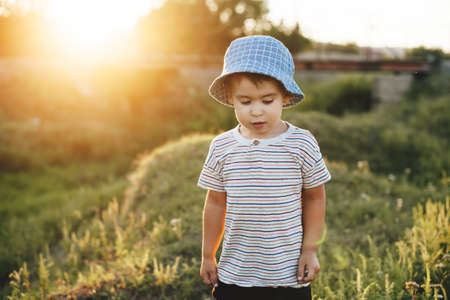 Small caucasian boy is walking in a green field wearing a summer hat during a warm sunny eveningの写真素材