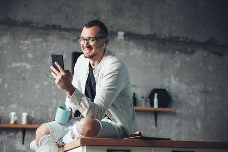 Caucasian businessman in white denim clothes is using a phone and earphones while drinking a coffee at homeの写真素材