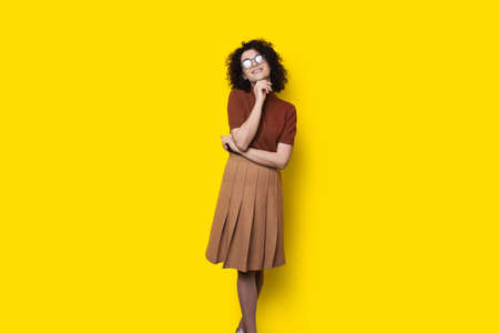 Lovely student with glasses and curly hair is posing on a yellow studio wall smiling at camera