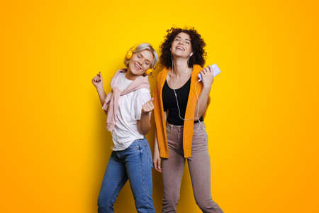Sisters listening music on yellow wall for mobile device design. Online lifestyle. Lovely girls dancing.の写真素材