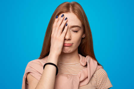 Woman with long red hair covering half of her face with the palm of her right hand. Portrait of a freckled young woman on a blue backgroundの写真素材