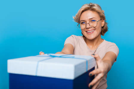 Caucasian woman handing out a birthday present. Portrait on a blue background. Smiling and looking at the cameraの写真素材