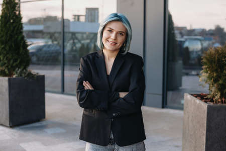 Portrait of a young blue-haired business woman standing in front of a company with folded hands. Smiling face.の写真素材