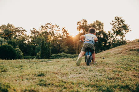 Back view of little kid in t-shirt riding bicycle alone in green grass field. Evening time. Sunsetの写真素材